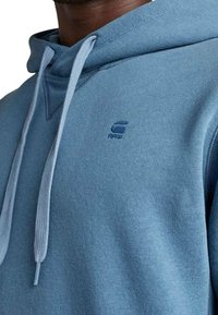 Sudadera azul hecha de tela suave, con un bolsillo canguro, capucha con cordón y un sutil logo bordado en el pecho.