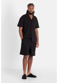 Chemise noire à manches courtes avec col et poche unique, assortie à un short noir. L'ensemble présente une coupe décontractée et une texture douce.