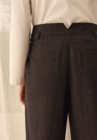 Braune, strukturierte Hose mit tailliertem Bund, zwei Gesäßtaschen und einem dezenten Glanz. Die Hand des Modells ruht seitlich, dazu ein helles Hemd.