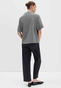 Frau mit einem Haarknoten, die ein graues kurzärmliges Strickoberteil, schwarze lockere Jeans und schwarze Ballerinas trägt, steht vor einem schlichten weißen Hintergrund.