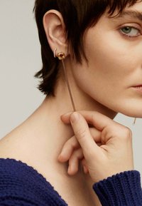 Pendientes de cadena dorada con una piedra preciosa cuadrada colorida y pequeños cristales. La mano de la modelo toca la cadena, mientras lleva un suéter texturizado en azul marino.