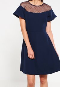 Laivastonsininen mekko, jossa on lyhyet kelluvat hihat ja läpinäkyvä pisteyksikkö. Sileä kangas, istuva vyötärö, liehuva hame ja polvimittainen leikkaus.