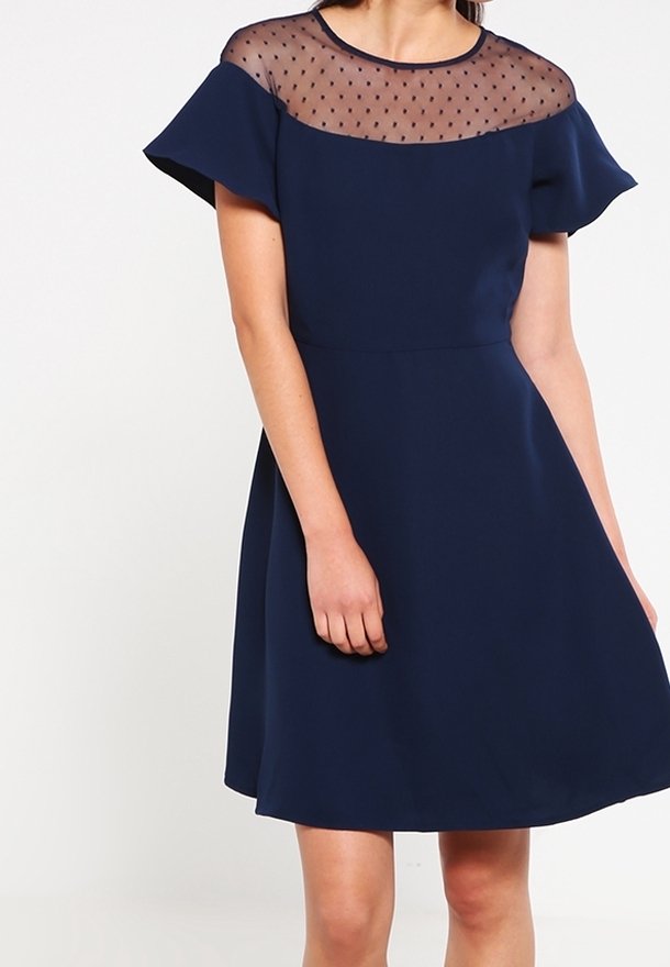 Laivastonsininen mekko, jossa on lyhyet kelluvat hihat ja läpinäkyvä pisteyksikkö. Sileä kangas, istuva vyötärö, liehuva hame ja polvimittainen leikkaus.