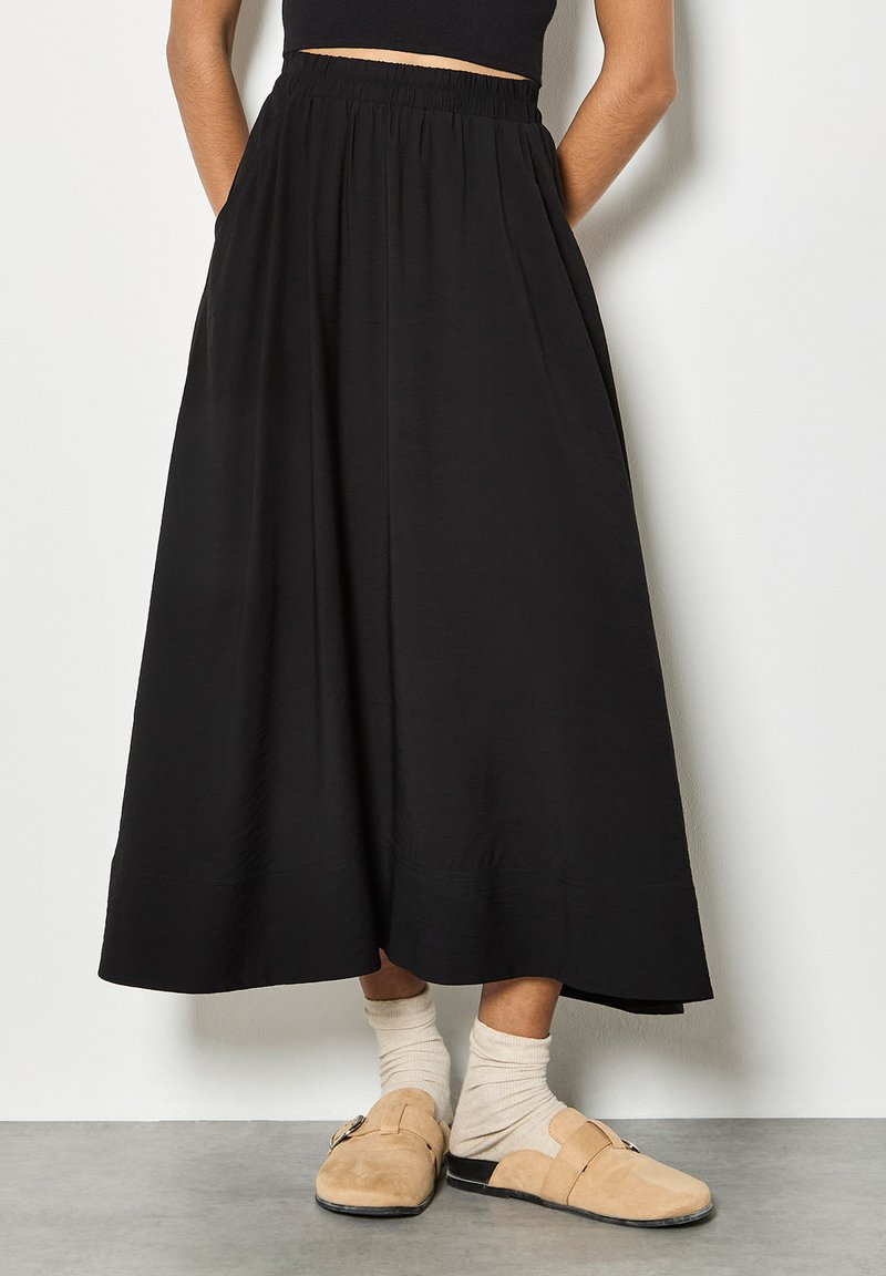 Personne portant une jupe midi noire, des chaussettes beiges et des chaussures plates en daim beige, debout contre un mur blanc.
