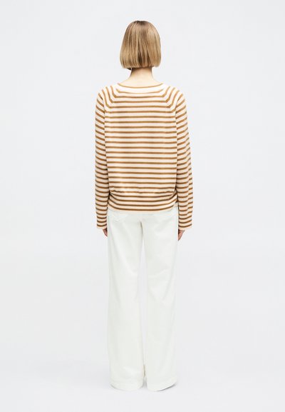 Rövid szőke hajjal rendelkező személy barna és fehér csíkos pulóvert és fehér bőszárú nadrágot visel, háttal áll fehér háttér előtt.