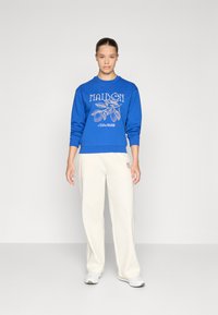 Felpa blu con stampa grafica, collo rotondo e maniche lunghe, abbinata a pantaloni ampi color crema. Sneakers bianche completano l'outfit.
