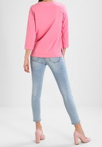 Haut rose à manches longues avec poignets retroussés, associé à un jean skinny bleu clair. Le mannequin porte des escarpins roses, mettant en valeur une tenue décontractée.