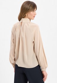 Blusa in morbido chiffon beige con colletto alto e schiena plissettata. Presenta maniche lunghe arricciate con polsini elasticizzati e un orlo curvo.