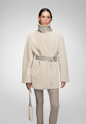 Beige nepbontjas met een hoge kraag, een ceintuur in de taille en zijzakken. Model draagt een kleine handtas met een bijpassende strap.