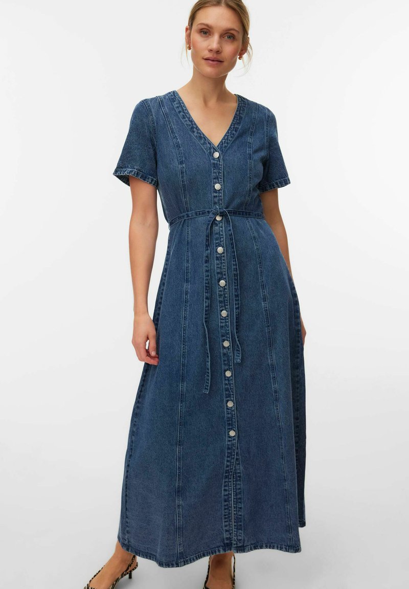 Naine kannab sinist nööpidega denimist maksikleit lühikeste varrukatega ja vööd, seisab ühevärvilise valge tausta ees.