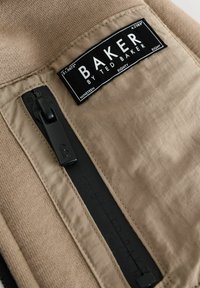 Beige kangas silt sisaldab "BAKER by Ted Baker", must tõmblukk "B" tõmbluku külge, ning tekstuuriga taust koos õmbluse detailidega.