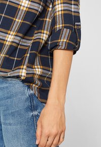 Námořnicky modrá kostkovaná košile se žlutými a bílými pruhy, s vyhrnutými rukávy a volným střihem, doplněná o roztrhané světle modré džíny.