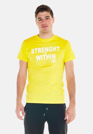 Jeune homme portant un t-shirt jaune vif avec le texte "STRENGHT IS WITHIN" et un pantalon noir, debout devant un fond blanc.