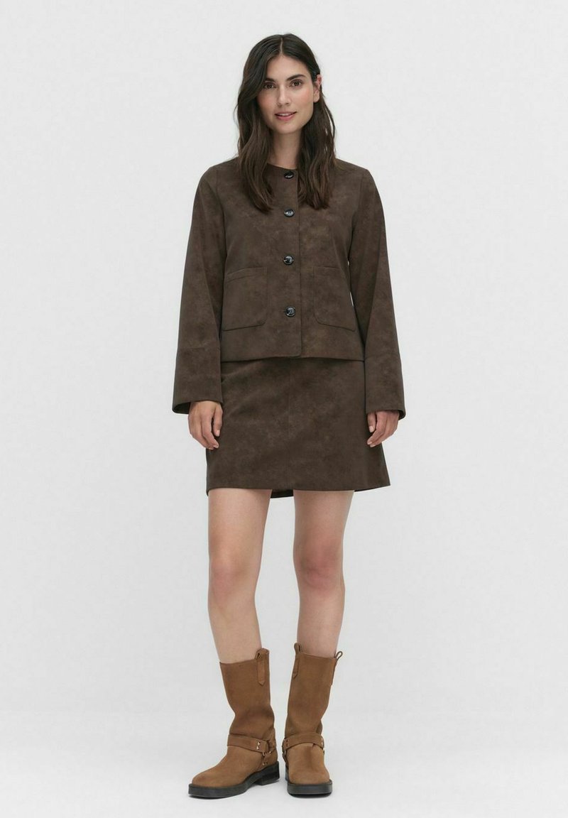 Femme portant une veste marron à boutons et une jupe assortie, avec des bottes marron mi-mollet, se tenant devant un fond clair.