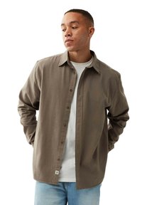 Brun button-up skjorte lavet af bomuld med en enkelt brystlomme, parret med en hvid t-shirt og lyseblå jeans. Glat tekstur, klassisk pasform.