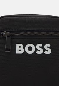 Borsa nera con texture, dotata di zip e lettering in rilievo bianco "BOSS". Il design è rettangolare con un aspetto elegante e moderno.