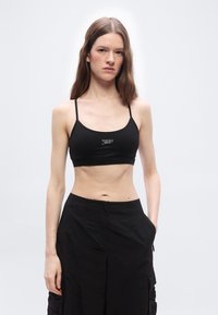 Jeune femme aux longs cheveux bruns portant un crop top noir à fines bretelles et un pantalon noir taille haute, debout devant un fond clair uni.