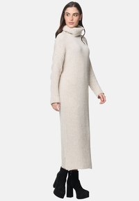 Beige geripptes Strickkleid mit hohem Kragen, langen Ärmeln und seitlichen Schlitzen, kombiniert mit schwarzen Plateaustiefeln.
