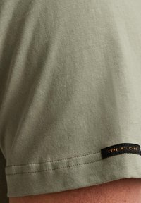 Olivegrünes Baumwoll-T-Shirt mit gerippter Textur und kurzen Ärmeln, mit einem schwarzen Etikett und orangefarbener Schrift am Saum.