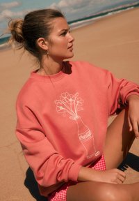 Korallefarbener Sweatshirt mit floralem Design und Coca-Cola Flasche Grafik. Weicher Stoff, lockere Passform und gerippte Bündchen. Wird über karierten Shorts getragen.
