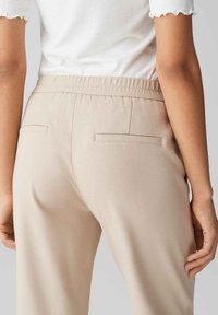 Beige broek met een gladde textuur, elastische tailleband en twee achterzakken; gedragen met een witte top met gegolfde zoom.