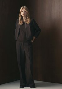 Giacca bomber nera sopra un maglione marrone scuro testurizzato, abbinata a pantaloni neri a gamba larga e scarpe scure, su uno sfondo scuro.