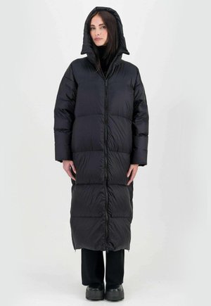 Zwarte gewatteerde jas met een capuchon, met een lange, doorgestikte uitstraling, een voorknofluiting en een oversized pasvorm, gecombineerd met donkere broek en schoenen.