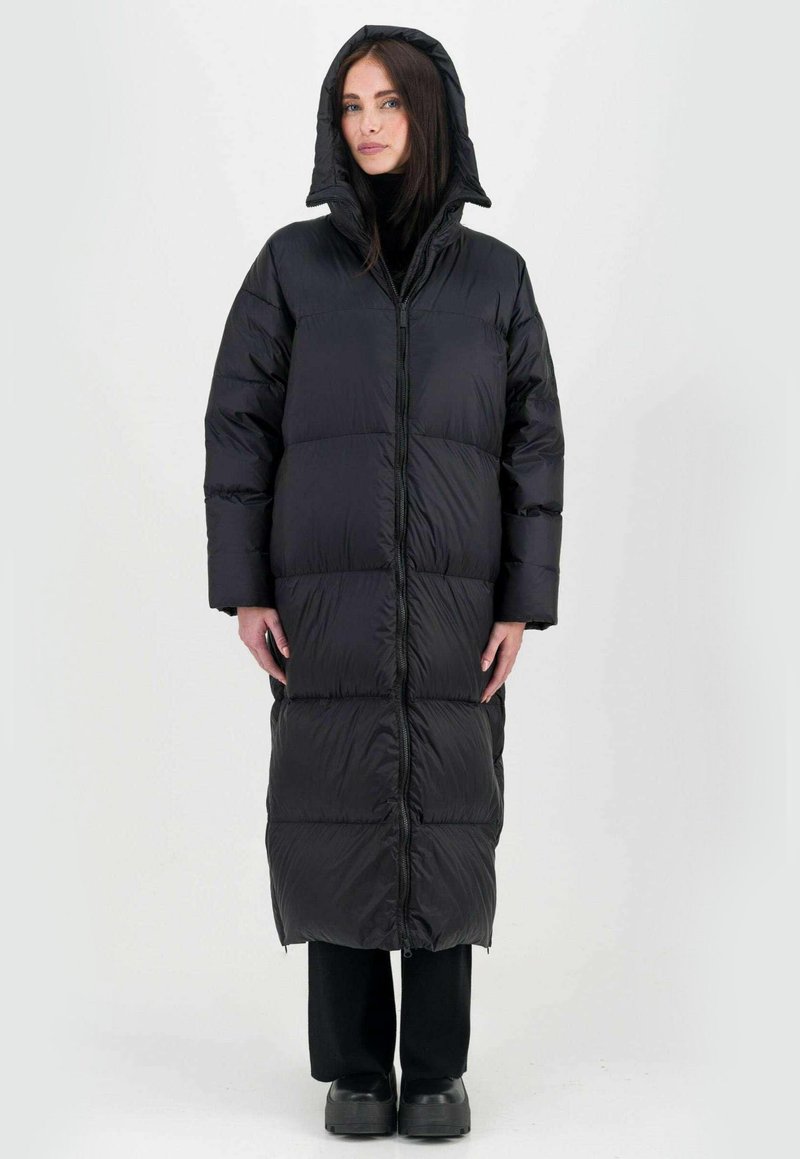 Doudoune noire avec capuche, à la coupe longue et matelassée, dotée d'une fermeture éclair à l'avant et d'une coupe oversize, associée à un pantalon et des chaussures sombres.