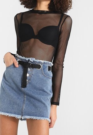 Mujer con top de manga larga de red negra sobre un sujetador negro, falda mini vaquera de talle alto con dobladillo deshilachado y cinturón de tela negro.