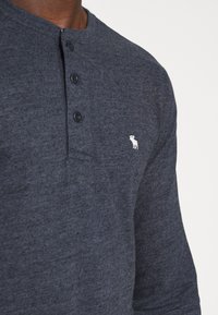 Tmavě modré dlouhé tričko s knoflíky vyrobené z jemné látky, se třemi černými knoflíky na límci a malým bílým logem na prsou.