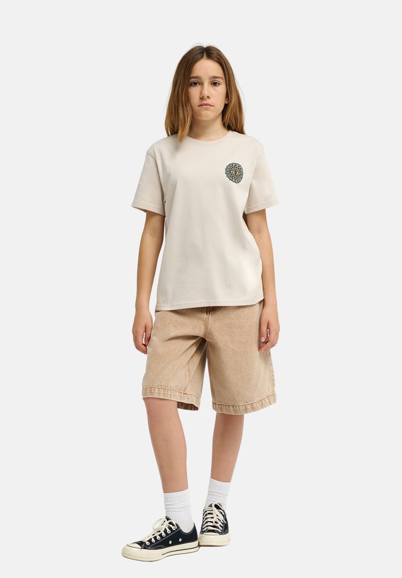 Camiseta de algodón beige con un parche gráfico redondo en el pecho, combinada con pantalones cortos de mezclilla marrón claro y zapatillas negras con suelas blancas.