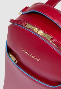 Rode leren rugzak met blauwe ritssluitingen. Voorzien van gouden hardware en een textuurafwerking. Logo prominent zichtbaar aan de voorkant.