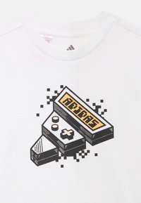 Valge puuvillane t-särk, millel on retro mängukonsooli graafika mustade joonte ja kollaste tekstidega, piksliliste detailidega.