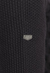 Schwarzer Strickstoff mit einem strukturierten Muster. Verfügt über ein silbernes "SD&CO"-Logo-Abzeichen in Form eines Wappens.