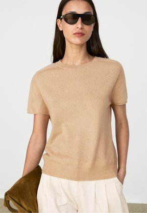 Femme portant des lunettes de soleil noires, un pull beige à manches courtes et un pantalon clair, tenant un sac souple marron dans une main.