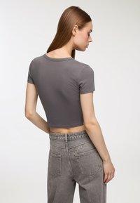 T-shirt grigia corta con maniche corte, realizzata in tessuto liscio. Il modello indossa jeans grigi sbiaditi a vita alta con cintura elastica e tasche.