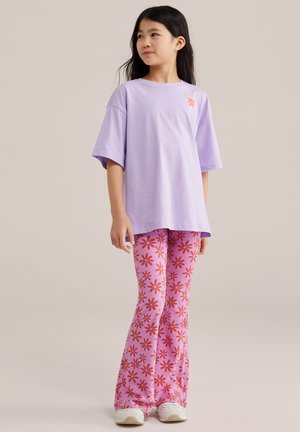 Pitkä mustahiuksinen tyttö pukeutuneena ylisuurkoon laventelinväriseen T-paitaan ja vaaleanpunaisiin kukkakuosisin levenevin housuihin valkoisilla tennareilla, seisomassa sivuttain.