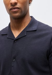 Mann med middels hudtone iført en mørk marineblå skjorte med krage, knappet foran, mot en ensfarget lys bakgrunn.