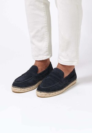 Zapatos náuticos de ante azul marino sin cordones, con puntera elevada, suela tejida de yute y detalle de costuras laterales, combinados con pantalones blancos remangados.