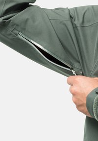 Manica di giacca verde con una tasca aperta, che mostra una superficie liscia e una chiusura con cerniera discreta. Il materiale appare leggero e funzionale.