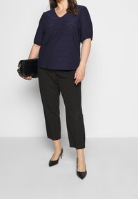 Námořnická modrá kratší rukávová blůza s výstřihem do V a detaily z průhledné krajky, v kombinaci s černými zkrácenými kalhotami a černými lodičkami s špičatou špičkou. Drží černou kabelku.