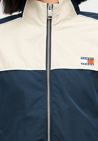 Μπουφάν με φερμουάρ σε ναυτικό μπλε και κρεμ αποχρώσεις, με γιακά που στέκεται όρθιος, αντίθετες βάτες στους ώμους και μικρό κεντητό λογότυπο στο στήθος.