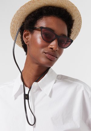 Donna con capelli corti e ricci che indossa un cappello di paglia con cinturino sotto il mento, occhiali da sole neri a forma di occhi di gatto e una camicia bianca con colletto.