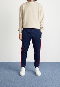 Beige sweatshirt med rund halsringning, tillsammans med navy blå träningsbyxor med röda sidosträck och en adidas-logotyp. Vita sneakers fulländar looken.