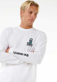 Camiseta de manga larga blanca hecha de algodón; presenta un logo en teal y negro. El texto en la manga dice "la búsqueda" en negro.