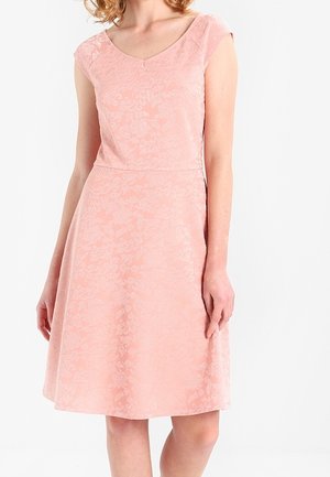 Femme portant une robe rose sans manches, longueur genou, avec un motif floral subtil et un décolleté en V, debout les mains détendues le long du corps.