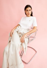 Witte katoenen t-shirt met een kleine geborduurde detail en beige wijde pantalon. Model zit op een chroomstoel tegen een roze achtergrond.