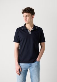 Camisa polo azul marino con cuello a rayas y pequeño logotipo en el pecho. La tela es suave y las mangas son cortas. Combinada con jeans de color azul claro.