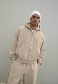 Bežna zip-up kapuca z ohlapnim krojem, ki ima velik kapuco in sprednji žep, v kompletu z usklajenimi bež hlačami. Nosite z belo pleteno kapo.