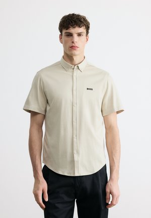 Jeune homme aux cheveux courts et bouclés portant une chemise beige à manches courtes avec des boutons et une étiquette "BOSS", ainsi qu'un pantalon noir, debout devant un fond blanc.